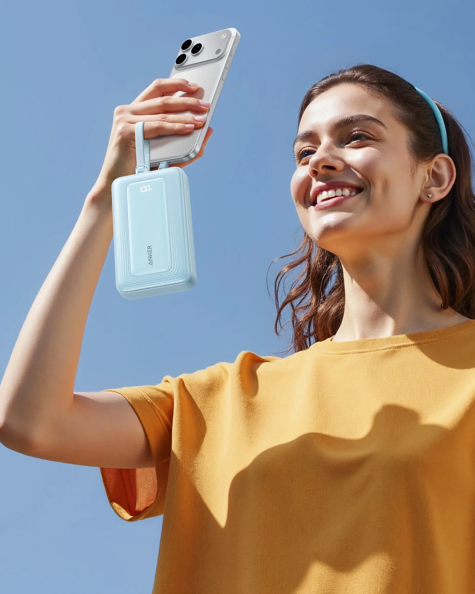 Junge Frau hält bei blauem Himmel ein Smartphone hoch, an dessen Ladebuchse die hellblaue Anker Zolo Powerbank mit 100 Prozent im Display hängt
