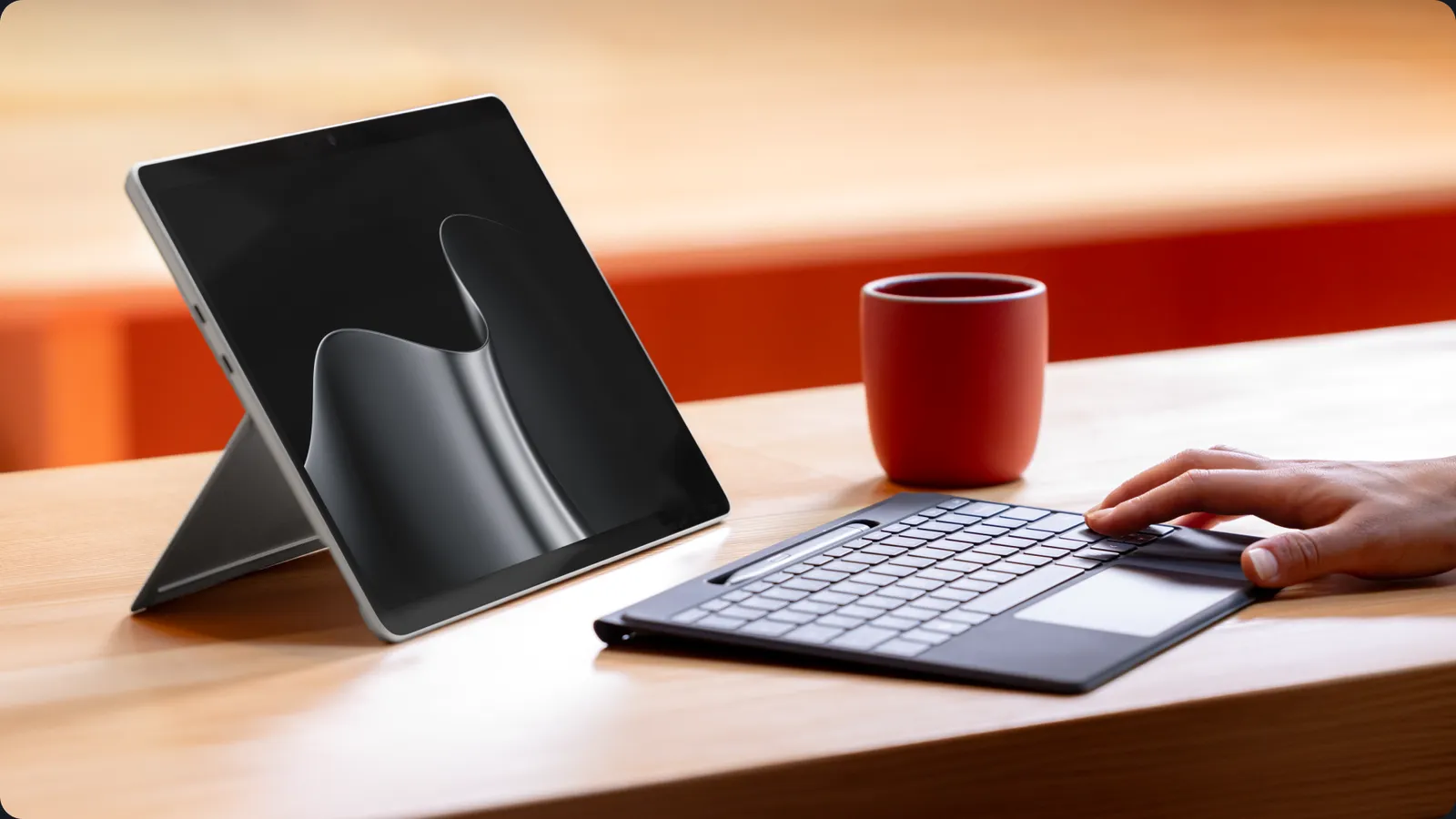 Surface Pro mit aufgestelltem Ständer und angesteckter Tastatur auf einem Holztisch, daneben eine rote Tasse, eine Hand liegt auf dem Touchpad