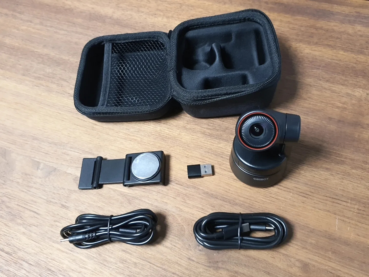 Lieferumfang der OBSBOT Tiny 4K: Kamera, Magnethalterung, USB-C-Kabel, Stromkabel und Aufbewahrungstasche auf einem Holztisch